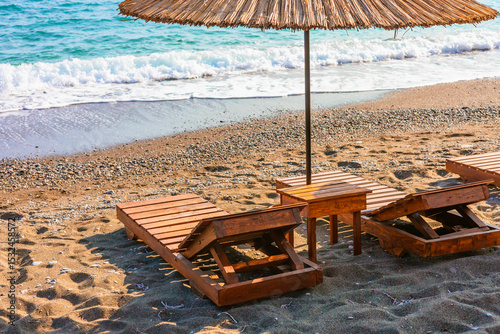 Fototapeta Naklejka Na Ścianę i Meble -  Wooden loungers are positioned in rows under straw umbrellas at a sea beach, inviting relaxation by the shore