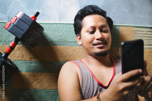 Man Relaxing After Exercise While Browsing Phone Beside Fitness Dumbbells