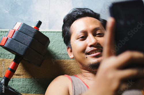 Man Relaxing After Exercise While Browsing Phone Beside Fitness Dumbbells