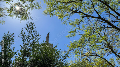 In luminous arboreal embrace, a solitary statue pierces azure skies, invoking Arbor Day and whispered tales of Verdant Reverie