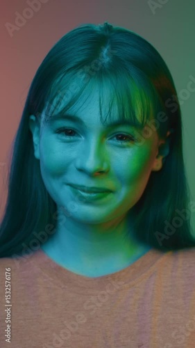 Happy Portrait of Young Woman Looking at Camera and Smiling in Neon Colored Lighting. Close Up View on Charming 20s Person with Shy Grin. Real Shot of Red Girl as Model Posing at Vivid Shooting Indoor