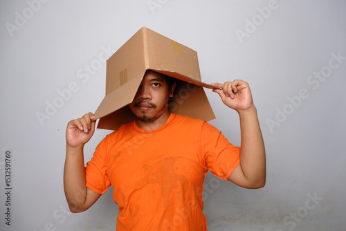 Young Asian man wearing an orange shirt with a cardboard box on his head, posing humorously against a plain background, symbolizing creativity and quirky expression.