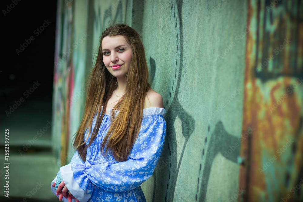 Fototapeta premium Young woman in blue dress posing near urban graffiti wall at night