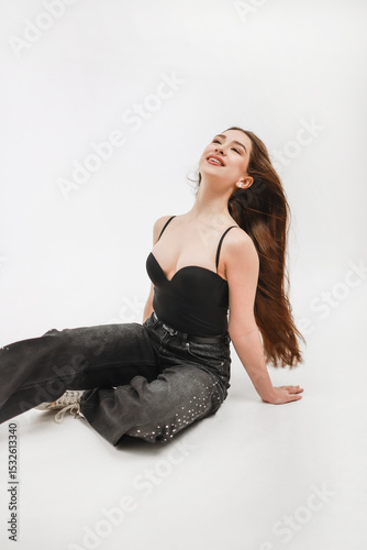 A girl in jeans and sneakers sits on a white background.