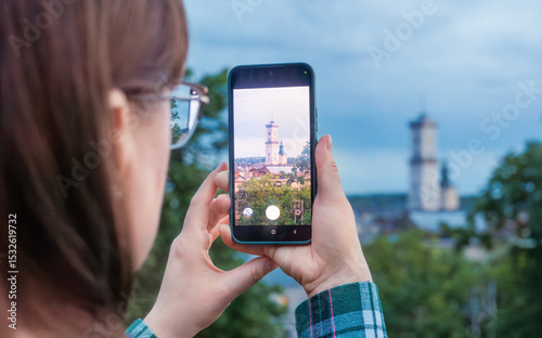 woman with long hair taking photo of cityscape using smartphone. Tourist capturing urban landscape in Lviv, Ukraine, with historic towers and trees. Travel, modern lifestyle, and digital technology 