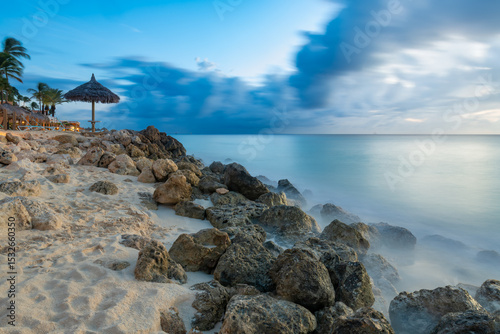 Tranquil coastline at twilight in Aruba, features motion-blurred waves washing over rocks beneath a blue and cloudy sky, with a beach palapa.