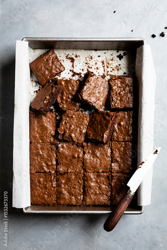 A baking tray filled with freshly cut chocolate brownies, accompanied by a knife on a parchment-lined surface.
