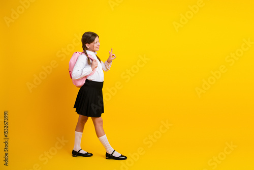 Confident young schoolgirl in uniform with a pink backpack posing on a yellow background, enjoying back-to-school season photo