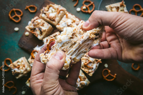 Overhead shot of hands pulling apart a gooey marshmallow rice treat with pretzels and white chocolate, with a wooden board pretzels and chocolate scattered around