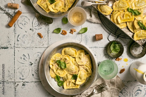 cooked ravioli in different pans with wine glasses and cork, basil leaves and nuts, olive oil on tiled table top, typical food from Liguria, Italy