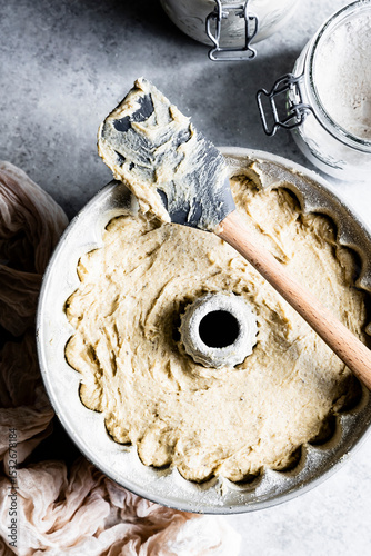 A bundt cake pan filled with cake batter, accompanied by a spatula and jars of flour on a textured surface.