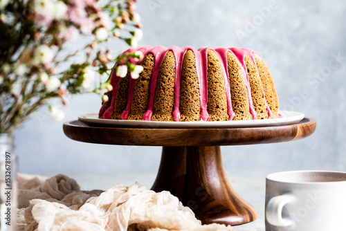 A bundt cake with pink icing is displayed on a wooden cake stand, with flowers and a cup nearby.