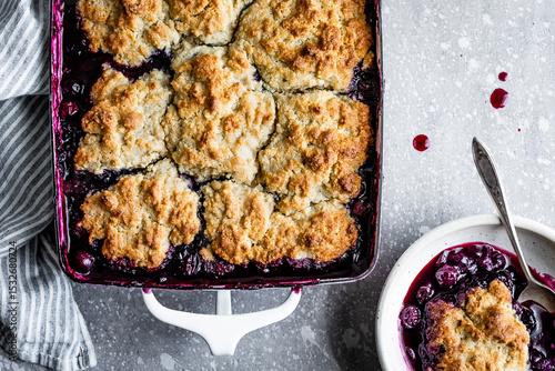 A freshly baked blueberry cobbler in a baking dish with a portion served in a bowl beside it.