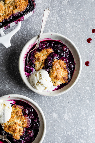 Two bowls of blueberry cobbler with vanilla ice cream on a speckled gray surface.