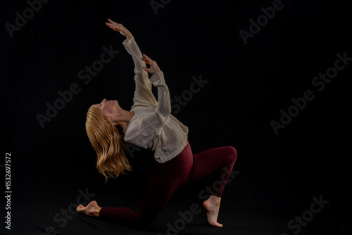 a person performing a yoga pose, balancing on one foot while holding the other with an opposite hand. They are dressed in a light-colored sweater and maroon leggings, standing against a dark backgroun