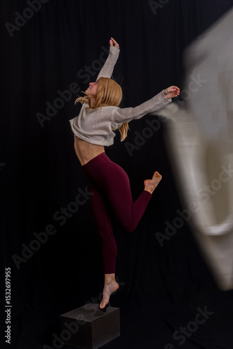 captures a dancer in a graceful pose on a black box against a dark backdrop. Dressed in a light gray sweater and maroon leggings, the dancer extends their arms upward while maintaining balance with on