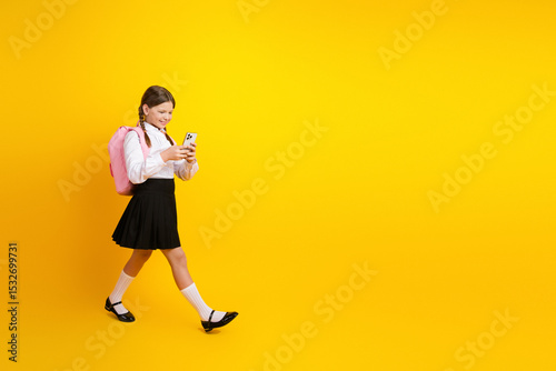 Happy schoolgirl in uniform with pink backpack using smartphone against vibrant yellow background photo