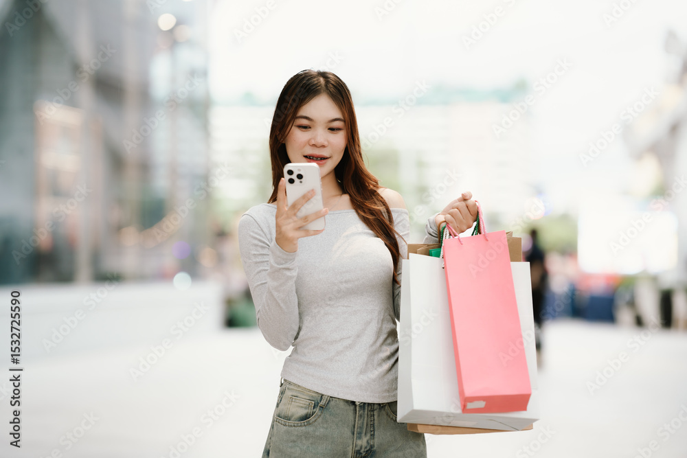 Fototapeta premium A young Asian woman with braces smiles while holding shopping bags and a smartphone outside a store.