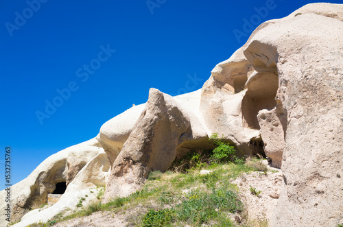 Natural rock formations with ancient cave dwellings carved into soft tuff rock in Cappadocia, Turkey. Scenic summer landscape of Cappadocia with arid terrain, bright blue sky and sparse vegetation.