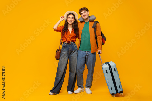 Happy European spouses ready for vacation, standing with suitcase, man wearing travel neck pillow and backpack, couple posing over yellow background
