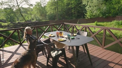 woman setting the table for breakfast on a terrace of a remote cabin in the woods with a lake view talking to her dogs