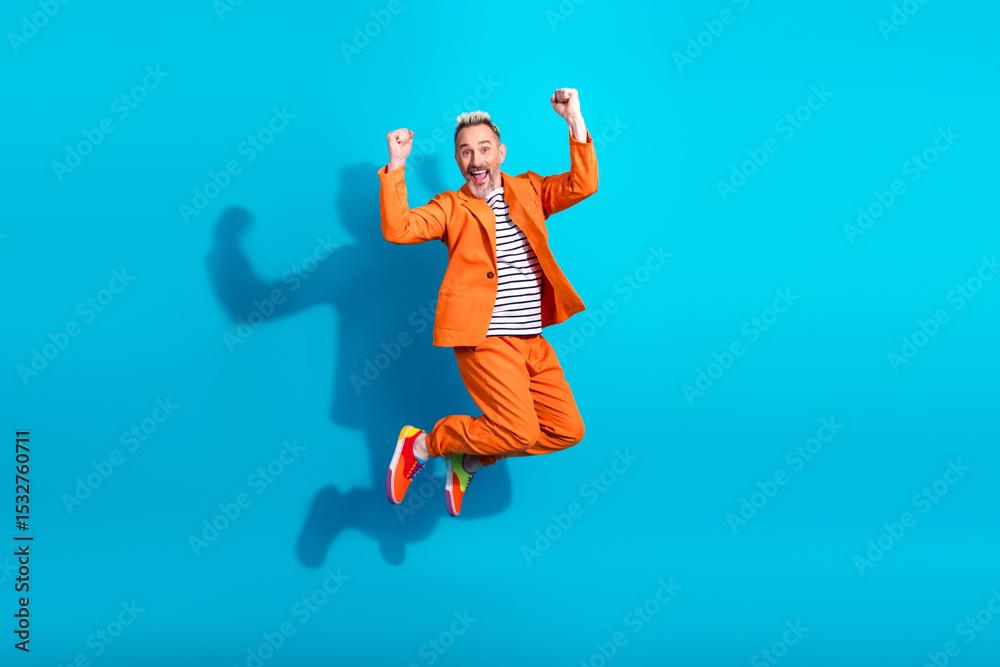 Obraz premium Charismatic gentleman in stylish orange outfit jumping with excitement against a vibrant blue background symbolizing joy