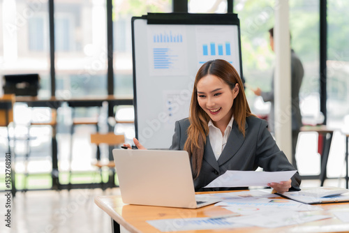 Beautiful Asian businesswoman working with laptop computer analyzing marketing data on screen while sitting at desk and working in accounting, finance and investment consulting.