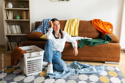 Tired woman feeling stressed over laundry chores, sitting on floor among dirty clothes with frustration, exhausted emotion at home