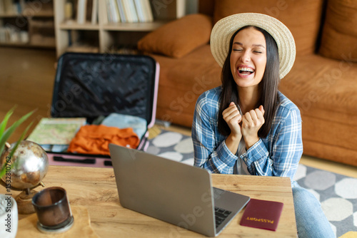 Cheap flight. Emotional happy lady gesturing, clenching fists, getting ready for journey, sitting in front of laptop with packed suitcase on background