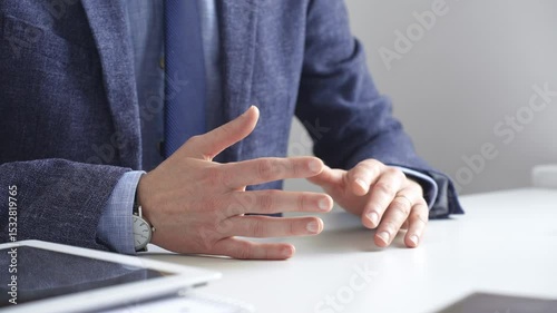 Professional businessman gesturing while discussing strategy, seated at white desk with tablet, eyeglasses, and wristwatch reflecting on sleek workspace surface. Business people concept