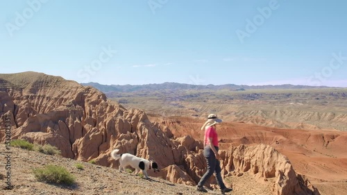 A woman and her dog, both hikers, descend from the mountains along the edge of a cliff above the red desert in the Boguty Mountains on a sunny spring day in Kazakhstan.