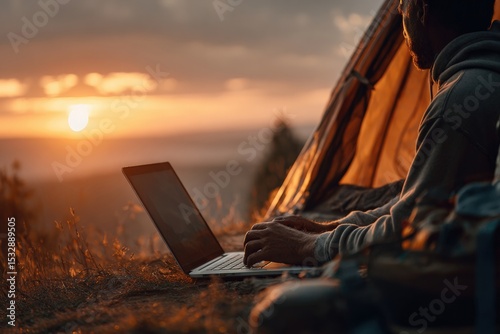 Digital nomad typing on laptop keyboard while sitting near tent in mountains at sunset, enjoying remote work and travel