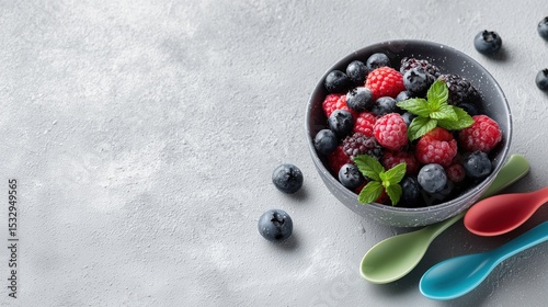 A bowl filled with ripe blueberries and raspberries is garnished with fresh mint leaves. Colorful spoons are placed beside, perfect for enjoying a delicious and nutritious treat