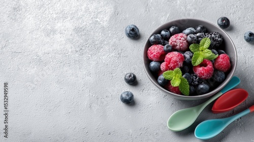 A bowl of fresh raspberries and blueberries is garnished with mint leaves. Colorful spoons surround the bowl on a soft gray surface, creating a vibrant, appealing arrangement