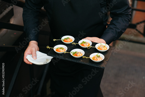 Waiter serving gourmet shrimp appetizers at event. A waiter in black uniform carries a tray of shrimp appetizers on skewers in small white bowls, ready for guests.