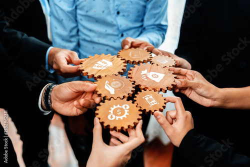 Hands of diverse professionals collaborate using wooden gears symbolizing teamwork and innovation in business strategy, aiming for reduced costs and enhanced success. Amity