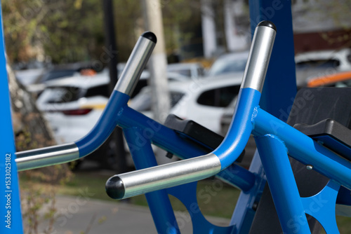 Close-up of blue outdoor gym equipment with metallic handles. Background features blurred cars in a parking area. Ideal for fitness and health concepts.