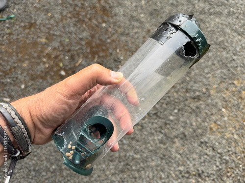 A hand can be seen holding a plastic bird feeder which has had holes chewed in it by a squirrel.