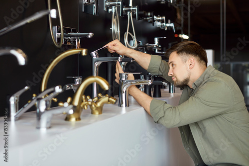 Fotografie Portrait of a young male customer choosing faucet for house standing in construction market or shop and examining plumbing
