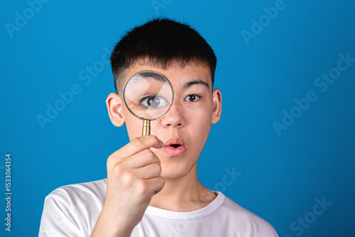 Portrait of a young asian surprised boy looking through a magnifying glass against blue background