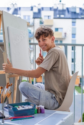 Cheerful, cute teenager at an easel learning to draw with colored paints and brushes on an easel, smiling sincerely while looking into the camera