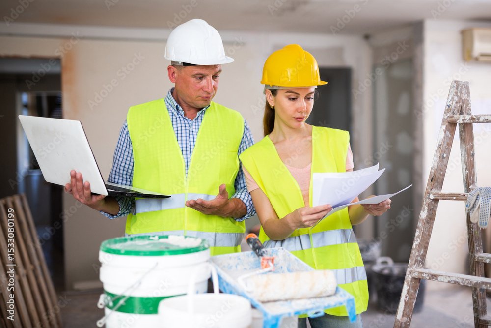 Fototapeta premium Portrait of woman architect and foreman holding blueprints and using laptop while discussing working process in house being renovated