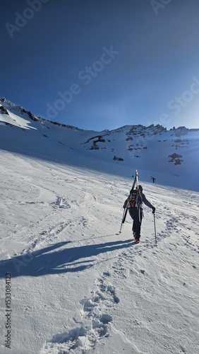 A person hiking up a snowy mountain with skis strapped to their back under a clear blue sky day