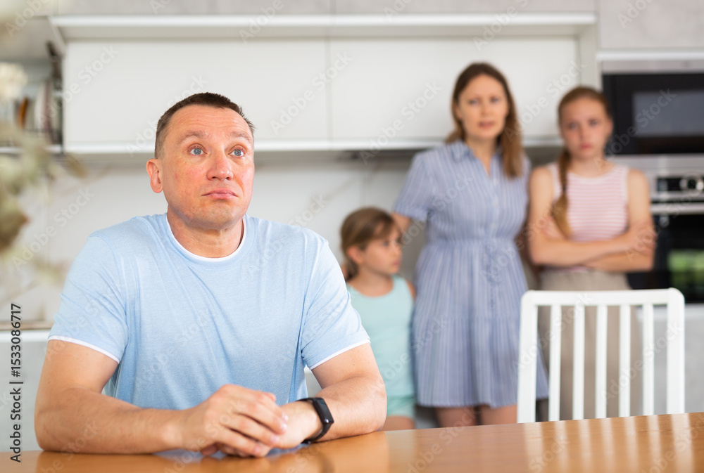 Fototapeta premium Upset father is sitting at the table, in background is a sad wife with small children. Family quarrel in home kitchen