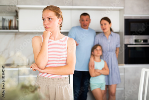 Portrait of upset teenage girl in home kitchen, frowning and crossing arms. Disgruntled parents reprimanding her while standing with younger daughter in blurred background. Emotional abuse concept