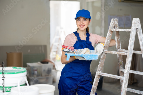 Confident young female house painter with work tools standing in house being renovated