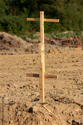 Simple wooden survey stake with crosspieces stands upright in dry, compacted soil, marking ground level or boundaries on a cleared construction site with brush and trees in the background.