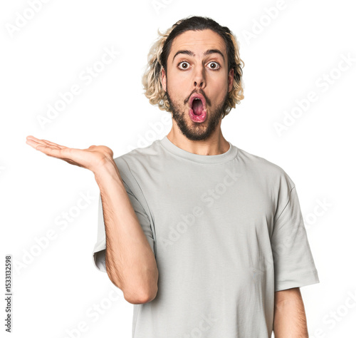 Young Caucasian man in studio setting impressed holding copy space on palm.