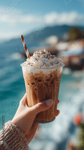 Hand holding refreshing iced coffee with whipped cream and chocolate powder topping, enjoying a sunny day by the sea