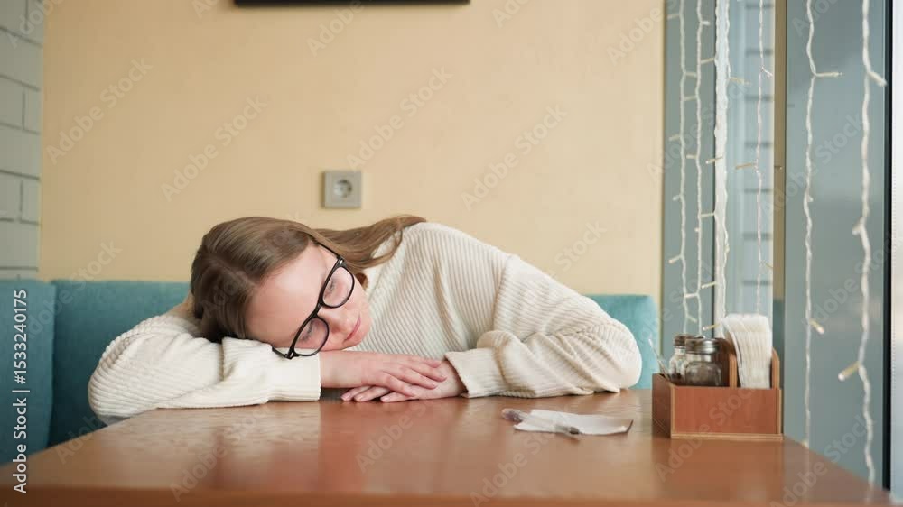 Student wearing glasses and cozy white sweater resting head on wooden table inside brightly lit room near window with fairy lights, suggesting tiredness